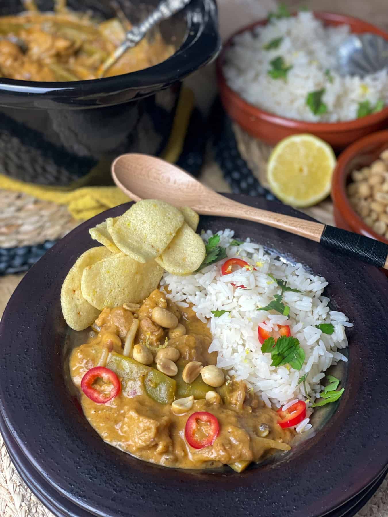 A close up bowl of Thai red curry with a wooden spoon on side of bowl, fresh lemon to the side with small bowl of rice and slow cooker pot in background.