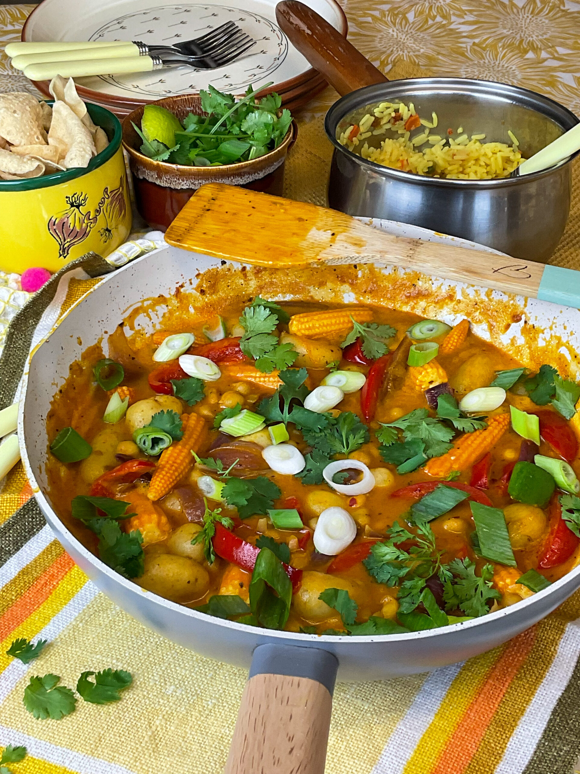 Mango chicken curry in pan with green garnishes, rice pan to side, bowl or poppadum's to side, yellow dinner mats.