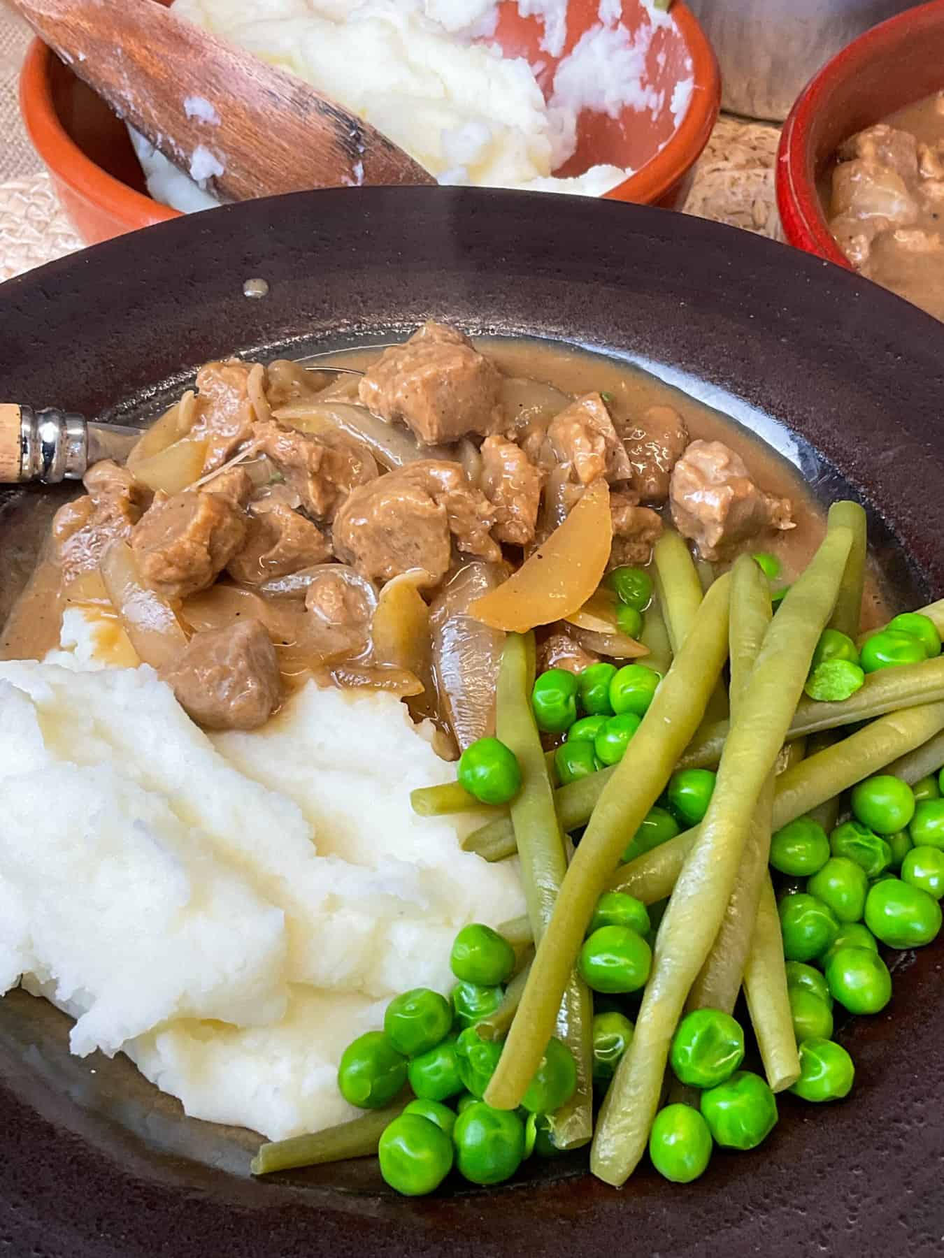 A close up of vegan Stroganoff in a brown bowl with mashed potatoes, green peas and beans to side, wooden handled spoon in bowl and a small bowl of mashed potatoes in background.