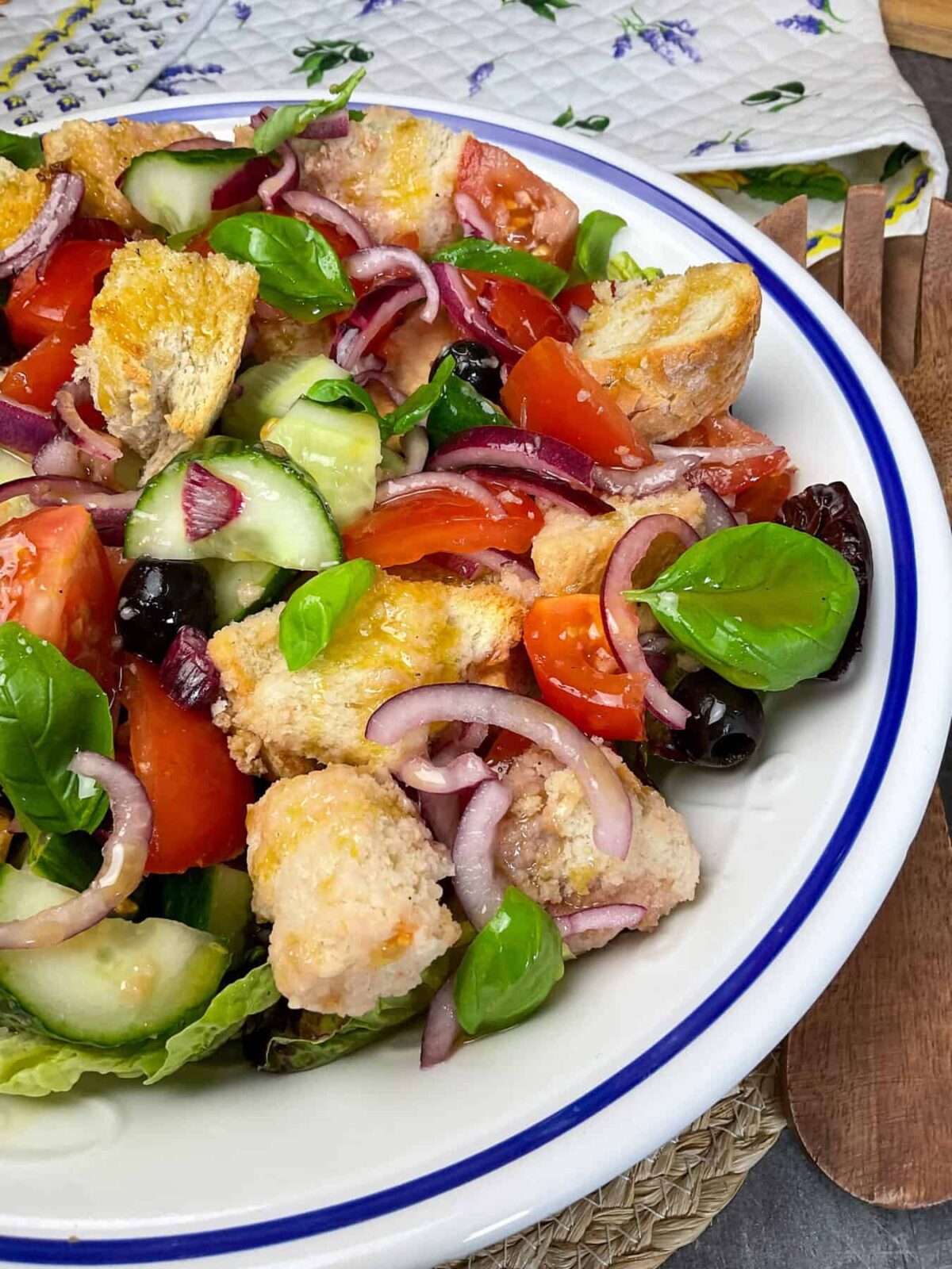 A side photo of Panzanella bowl of salad, with wooden salad spoon to side, and tea towel with flowers to background.