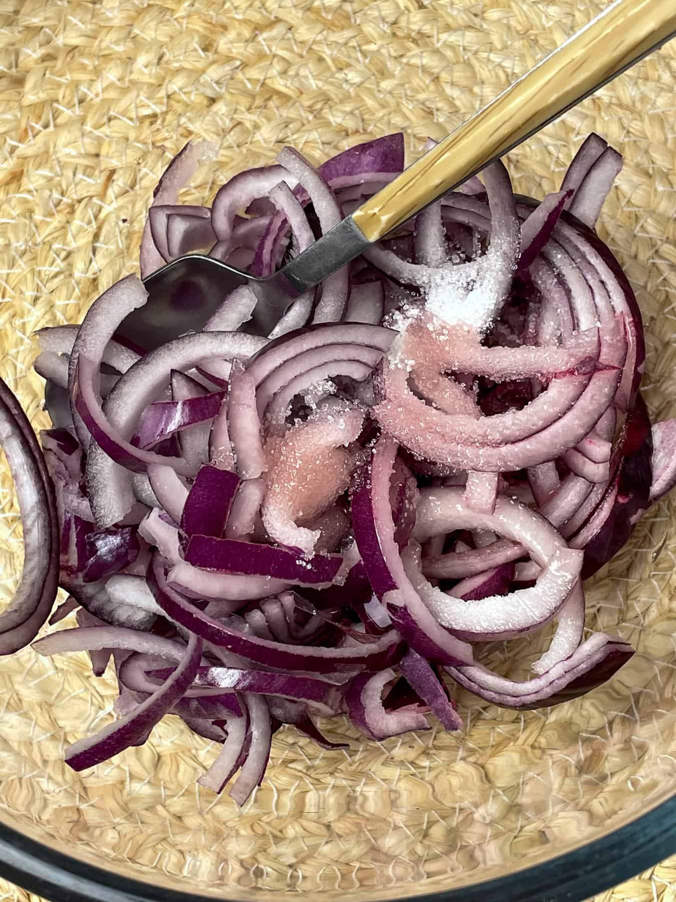 Red onion slices in glass bowl pickling in sugar and vinegar with spoon mixing.