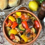 A brown bowl filled with ratatouille on a table with a crochet tablecloth, brown pepper shaker to side, bread rolls to side and lemon and raw whole vegetables in background.