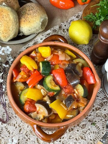 A brown bowl filled with ratatouille on a table with a crochet tablecloth, brown pepper shaker to side, bread rolls to side and lemon and raw whole vegetables in background.