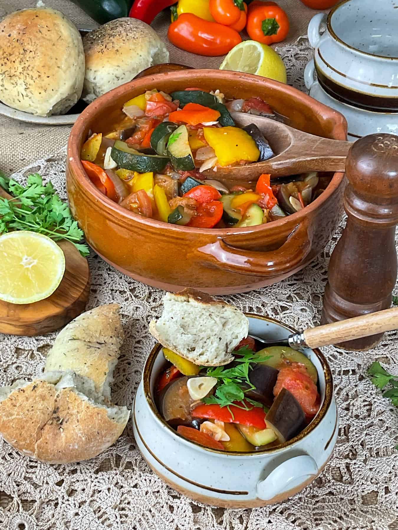 A table setting with small bowl of ratatouille and larger serving bowl in background, bread rolls to side, lemon wedge to side and empty bowls in background.