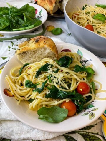 A bowl of garlic oil spaghetti with tomatoes and spinach and wedge of focaccia to side, larger pot to side, and bowl of fresh spinach to side.