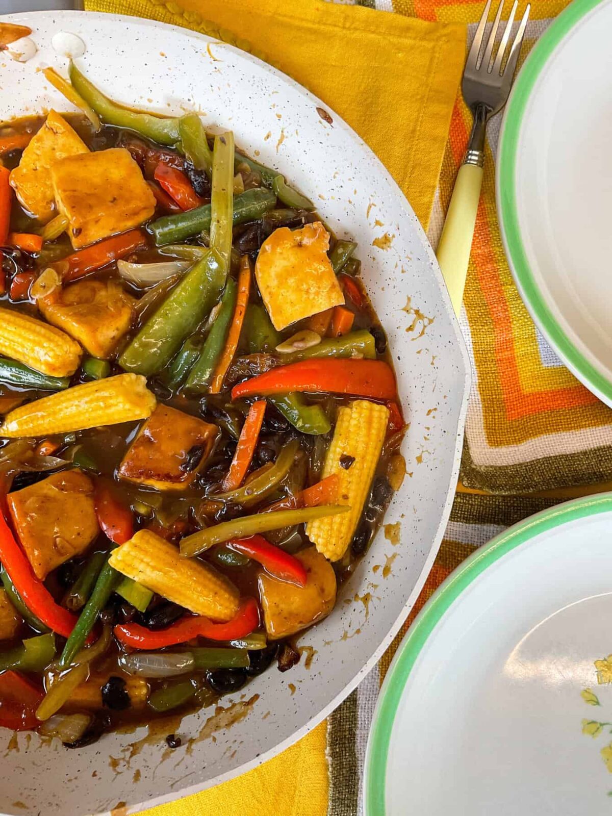 A side view of wok filled with sweet and sour tofu and veggies, with two green rimmed dinner plates to side and orange, brown and yellow striped table mats and yellow handled cutlery forks.