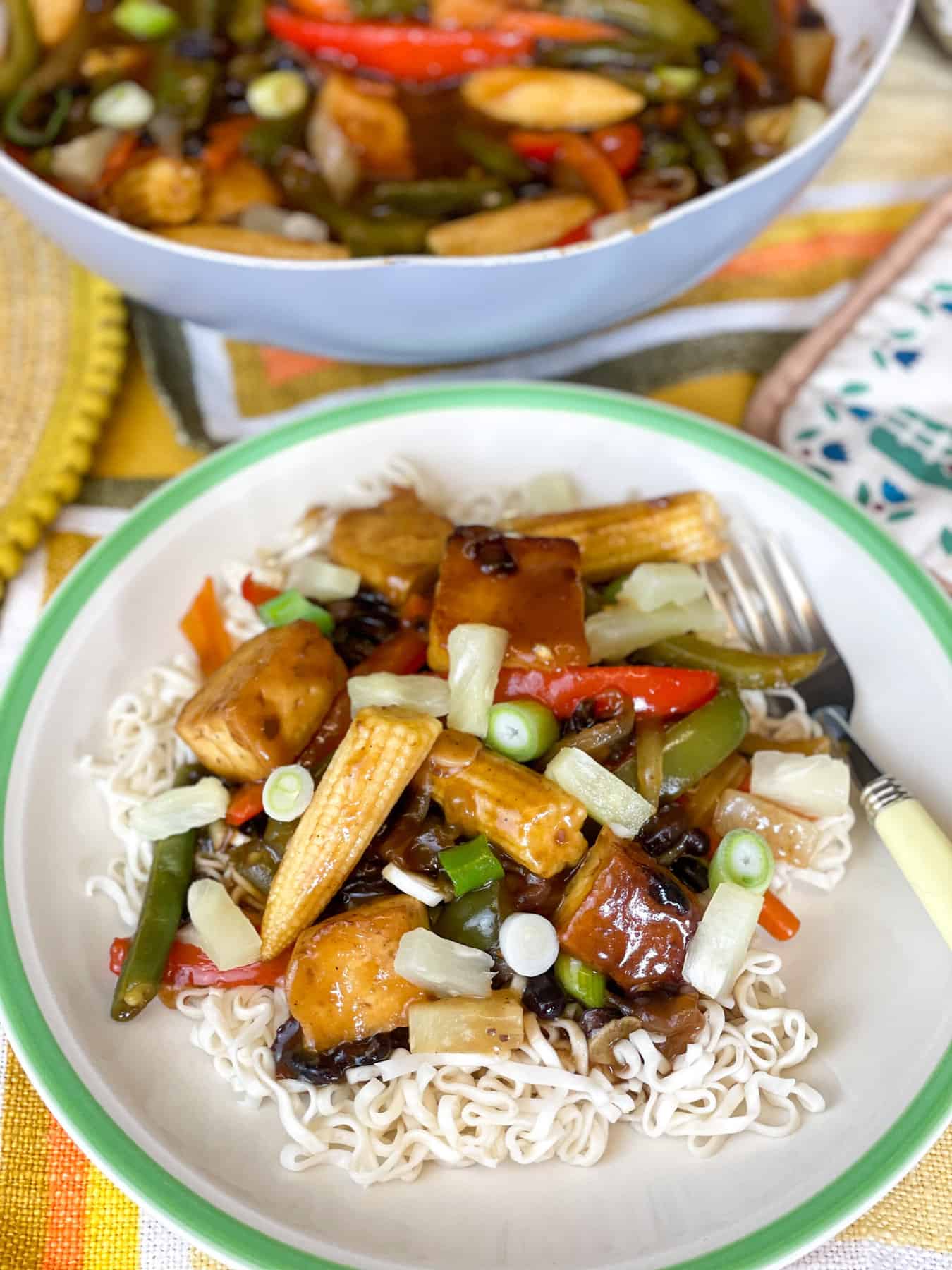 Sweet and sour tofu, veggies and black beans served over noodles on a green rimmed plate with pineapple pieces on top and sliced spring onions, wok in background.