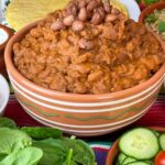 Vegan refried beans in a tan coloured serving bowl with white and green stripes, salad at the front, and tortilla wraps at the back, stripy colourful table mat background.