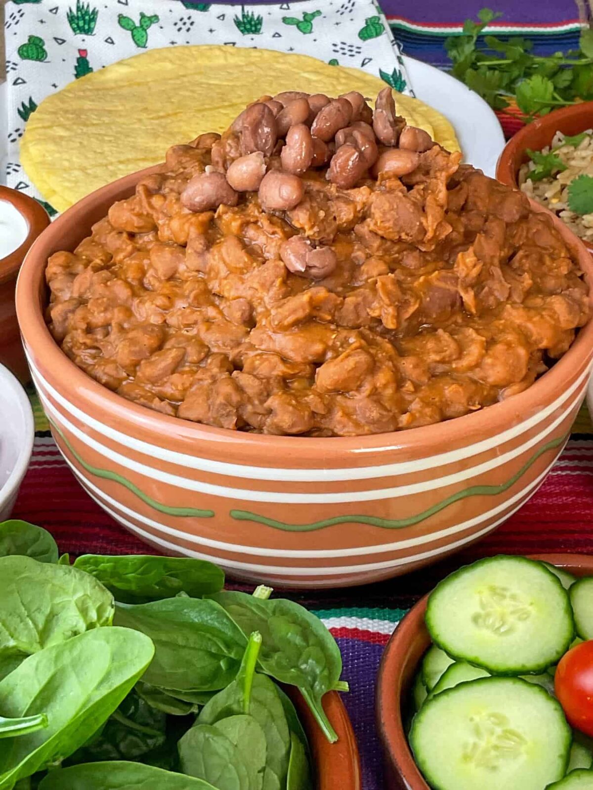 Vegan refried beans in a tan coloured serving bowl with white and green stripes, salad at the front, and tortilla wraps at the back, stripy colourful table mat background.