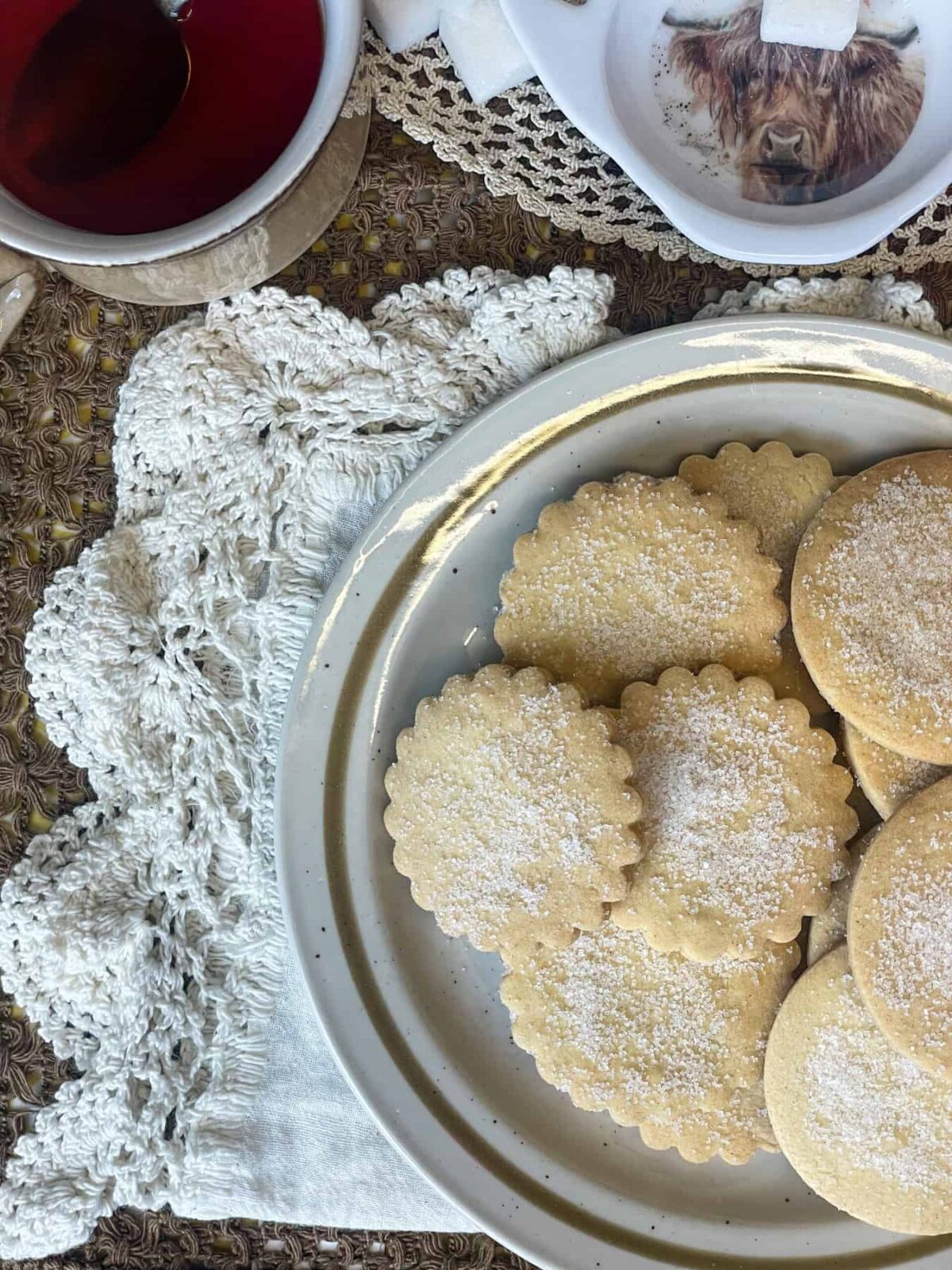 A side view of a plate of shortbread biscuits on an old-fashioned linen table cloth, cup of tea to side and small tea plate with image of Highland cow and sugar cubes.