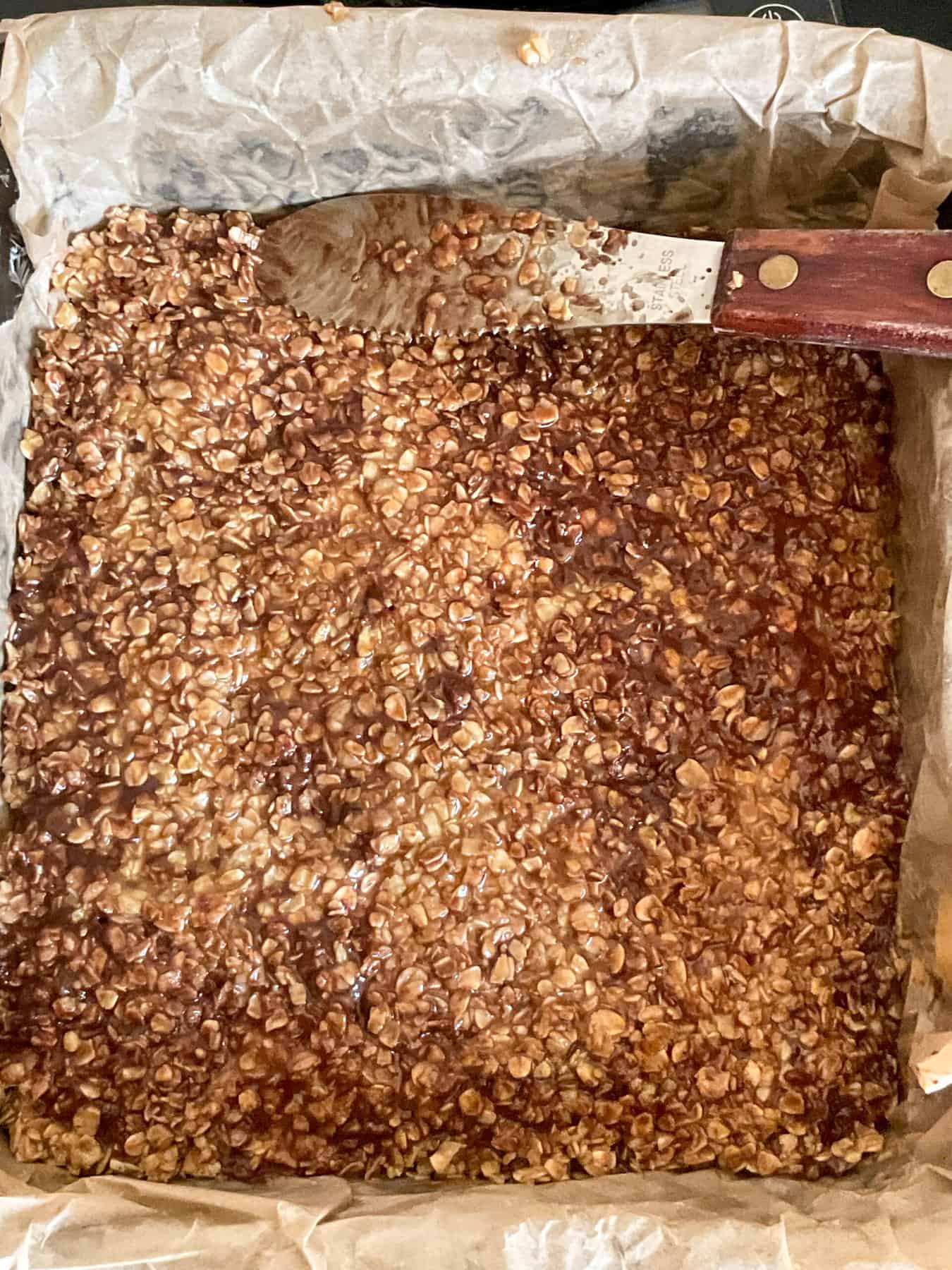 Chocolate flapjack mixture pressed into baking tray with a small wooden handled spatula.