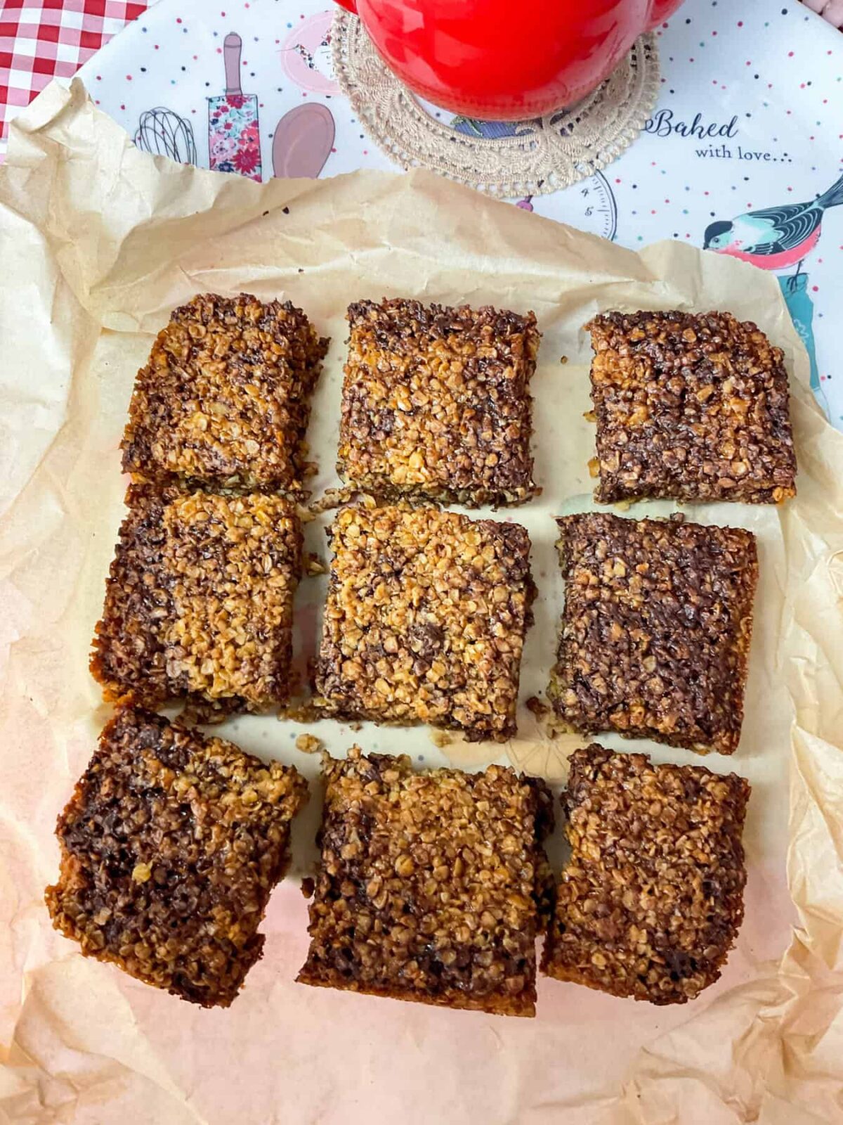 A batch of chocolate flapjacks sliced into 9 pieces on scrunched up baking paper, tea tray underneath and glimpse of a red teapot in background, red and white check table cloth.