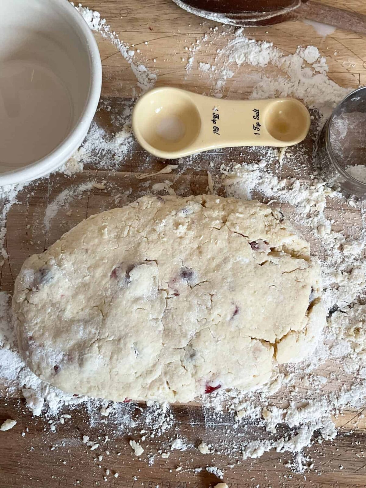 Scone mixture brought into a dough and sitting on a floured board with ceramic cream coloured tablespoon measurer and mixing bowl to side.