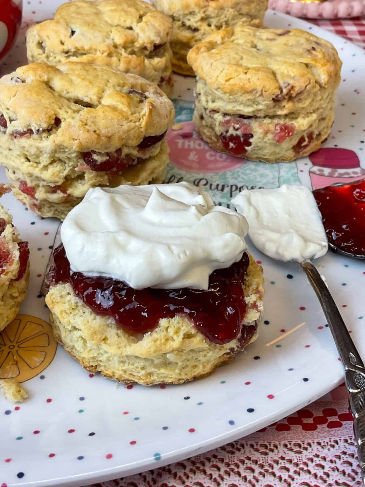 A tray of cherry scones with one halved and jam and cream added, cream and jam spoons to side.