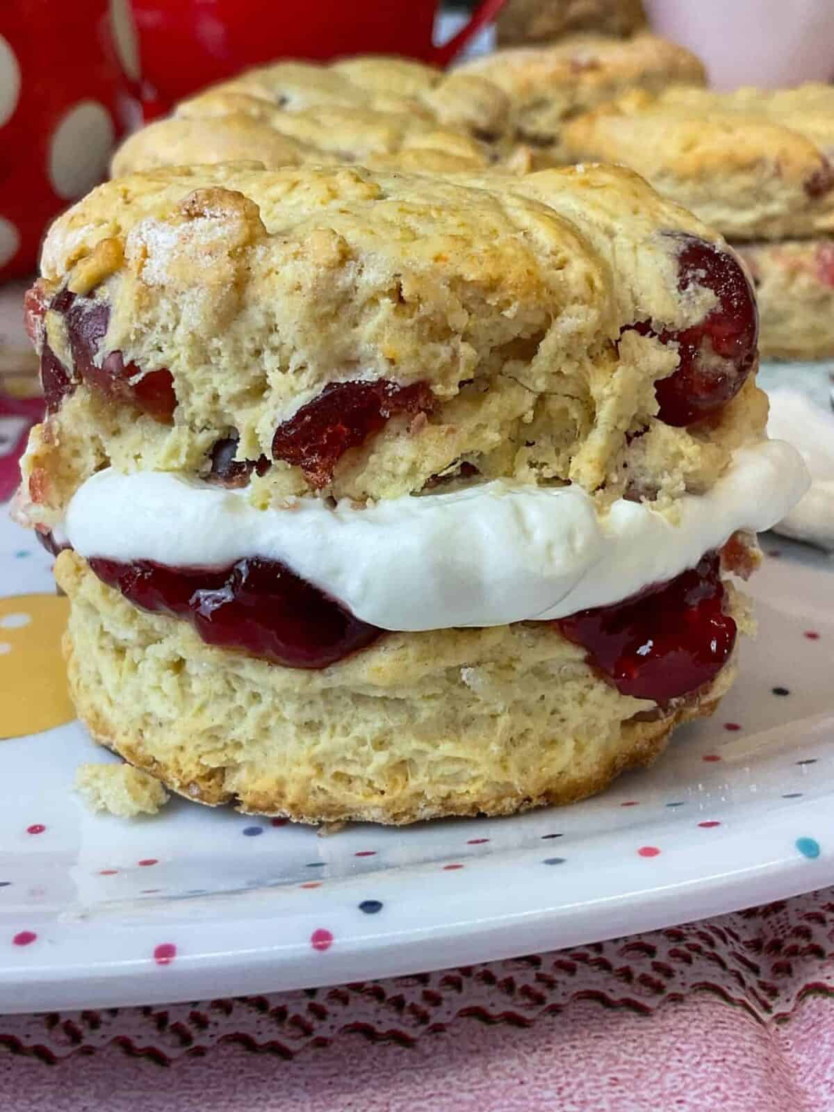 A close up of a cherry scone filled with jam and cream sitting on a spotty plate with pink mat underneath.