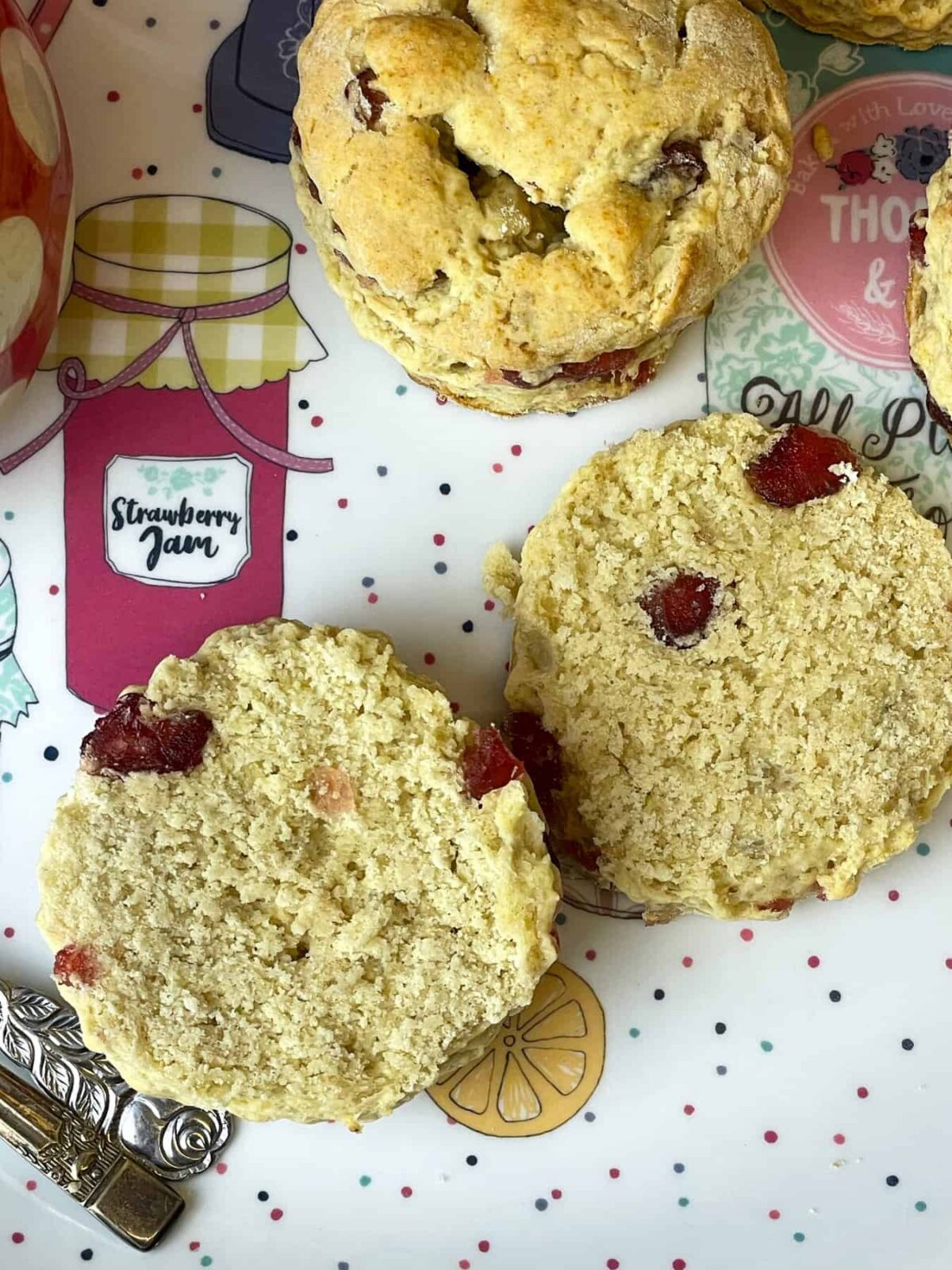 A cherry scone on a tea tray with images of jam pots and a speckled pattern, the scone is halved and a whole scone is in the background.