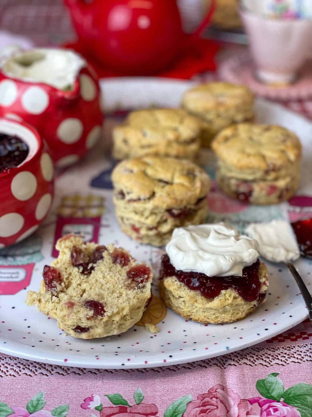 A cream tea tray with red teapot in background with pink cup, red spotty milk jug to side and matching jam bowl, scones on try with sliced scone to front, pink flower tablecloth.