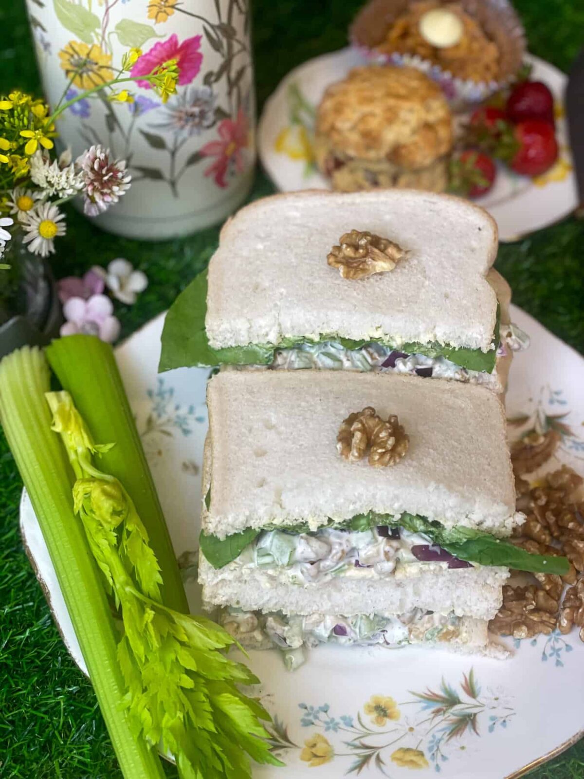 A plate of sandwiches with 3 celery stalks to the side and a flower flask and green grass background.