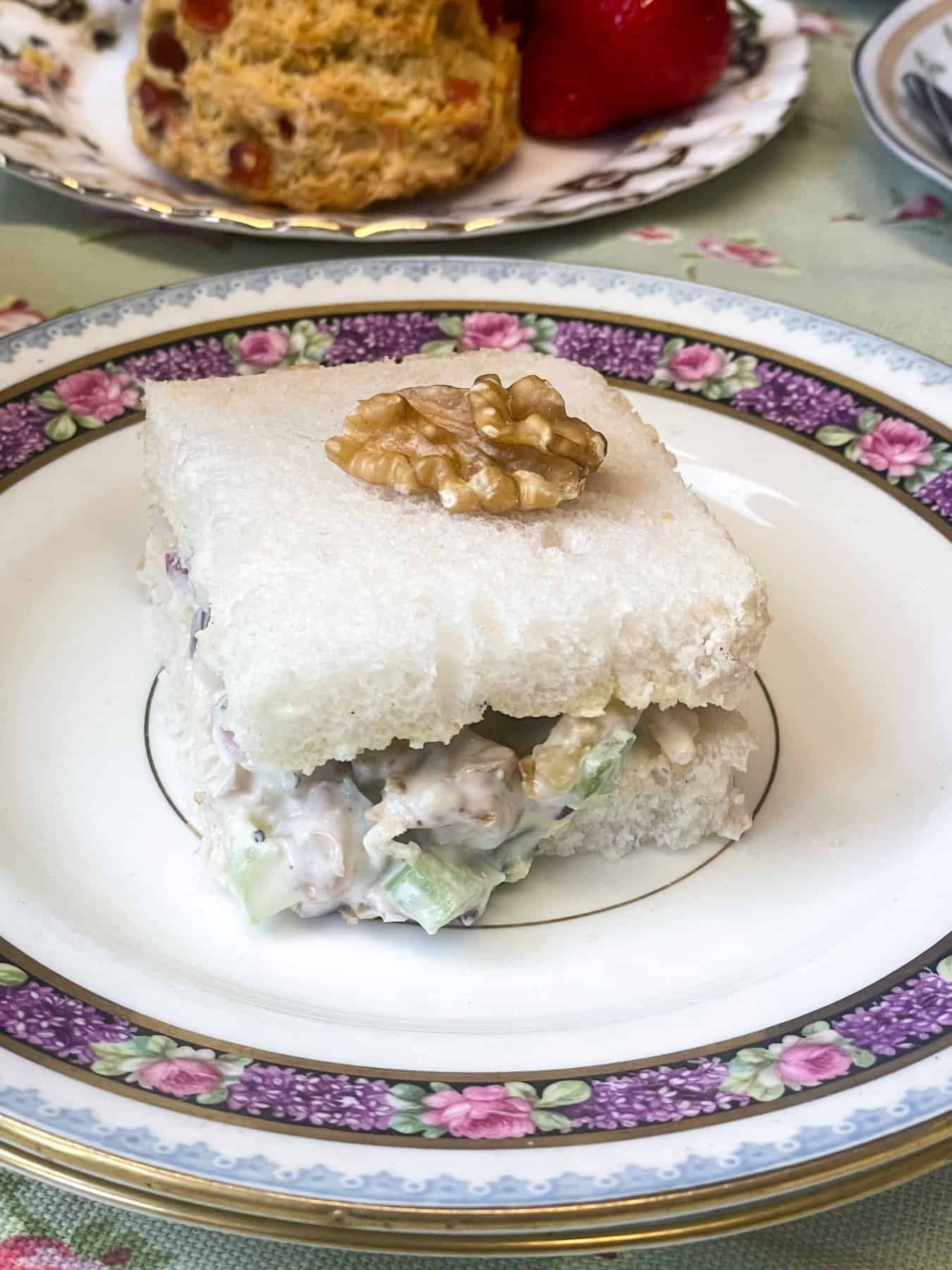 A small square sandwich on a purple flower rimmed plate, with a walnut garnish on top, and a plate in the distance with scones and strawberries.