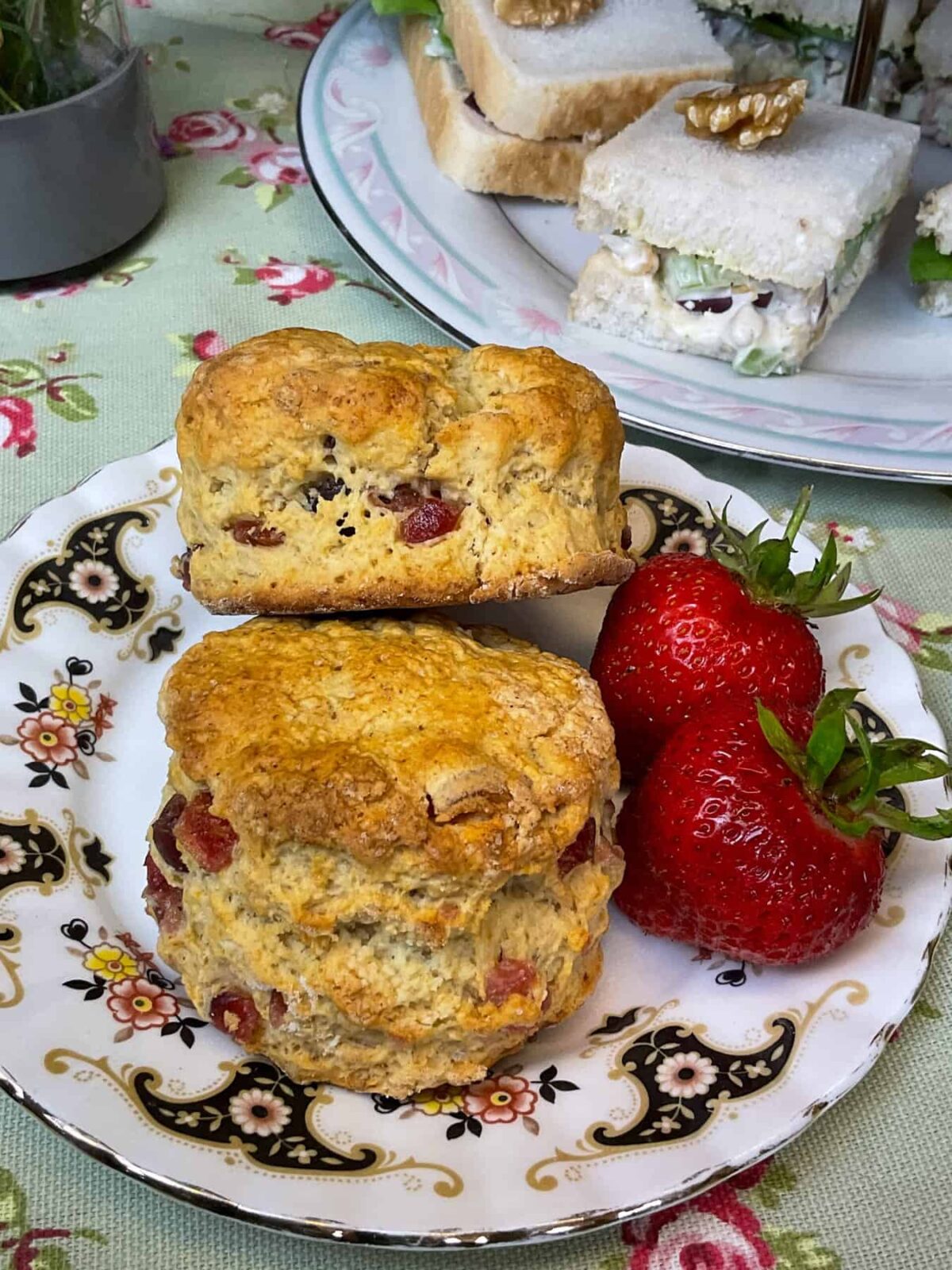 A vintage patterned plate with two cherry scones and two fresh strawberries, cake tier in background with dainty sandwiches, and a green tablecloth with flower images.