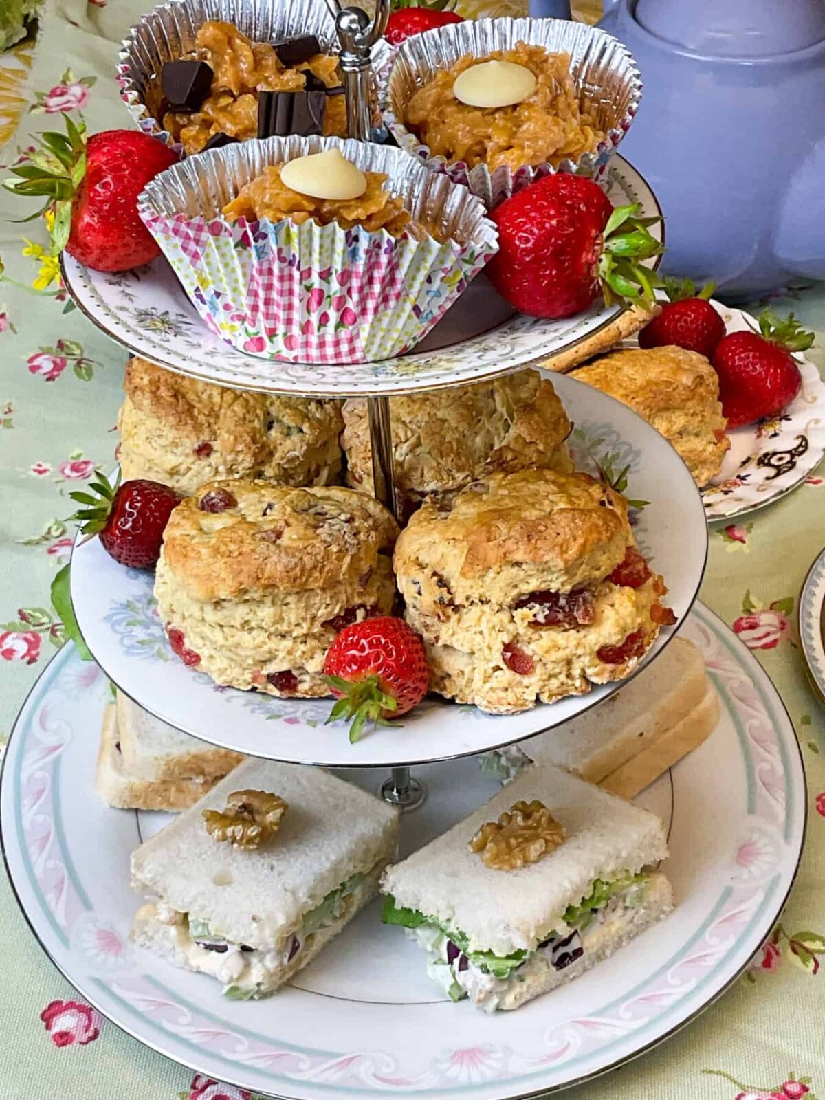 An afternoon tea tier with sandwiches at the bottom, cherry scones in the middle and peanut butter cornflake cakes at the top, strawberries decorating the layers, purple teapot in background and a green tablecloth with little pink flowers.