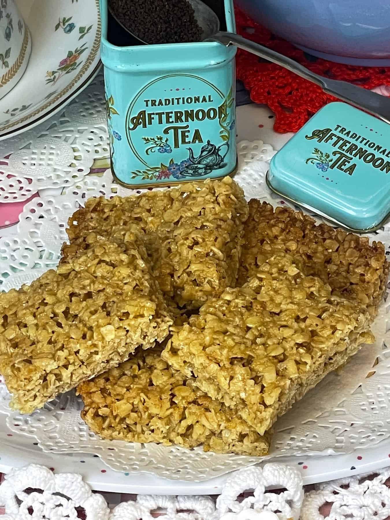 A plate of flapjacks on a paper doily, small tin of afternoon tea leaves in background with a tea spoon, teapot and cup in background.