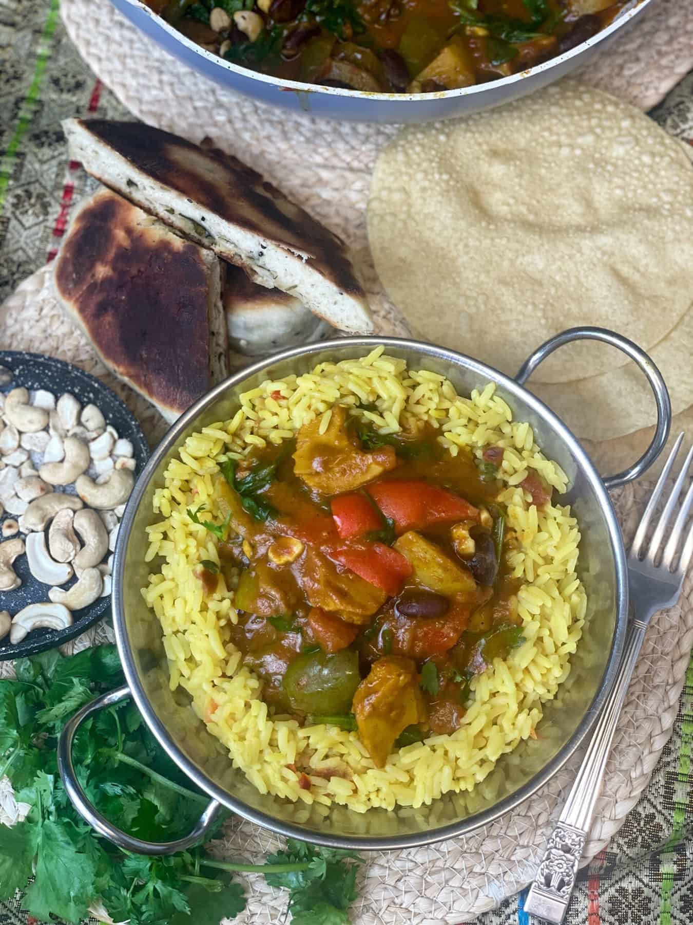 A single bowl of jalfrezi curry served with yellow rice, fresh coriander to the side, naan bread and poppadum's to the background along with Wok with curry, small skillet pan with toasted cashews to the side.