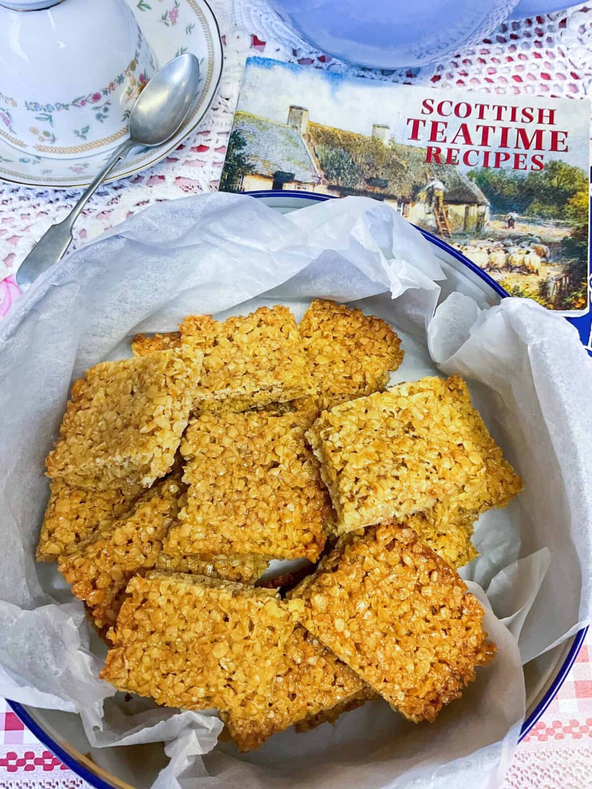 A biscuit tin filled with flapjack pieces, Scottish teatime small book to side, lilac teapot in background with vintage tea cup, saucer and spoon.