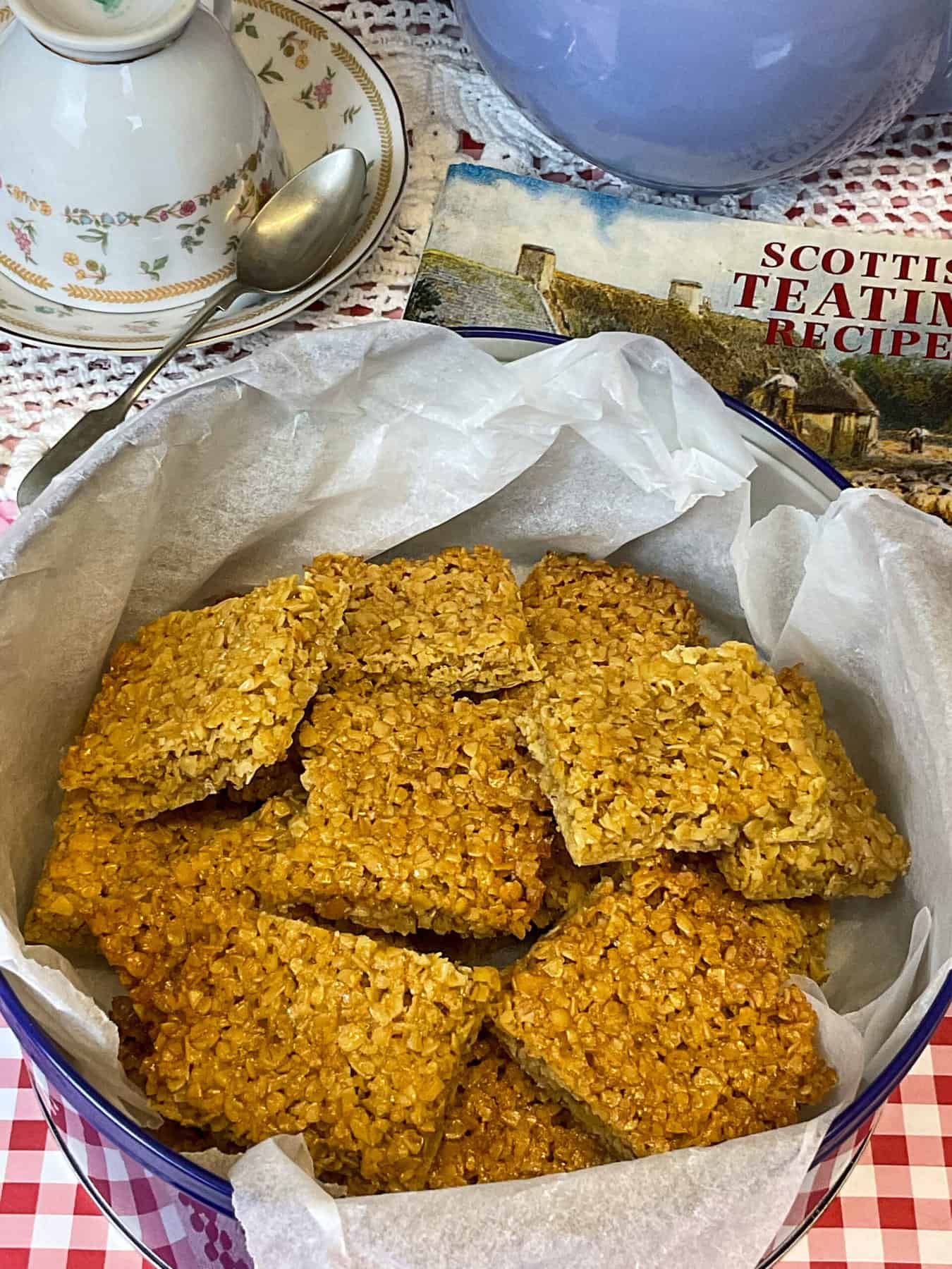 A blue biscuit tin filled with flapjack squares, purple tea pot to side with up turned cup and saucer, and tea spoon, red check table cloth background.