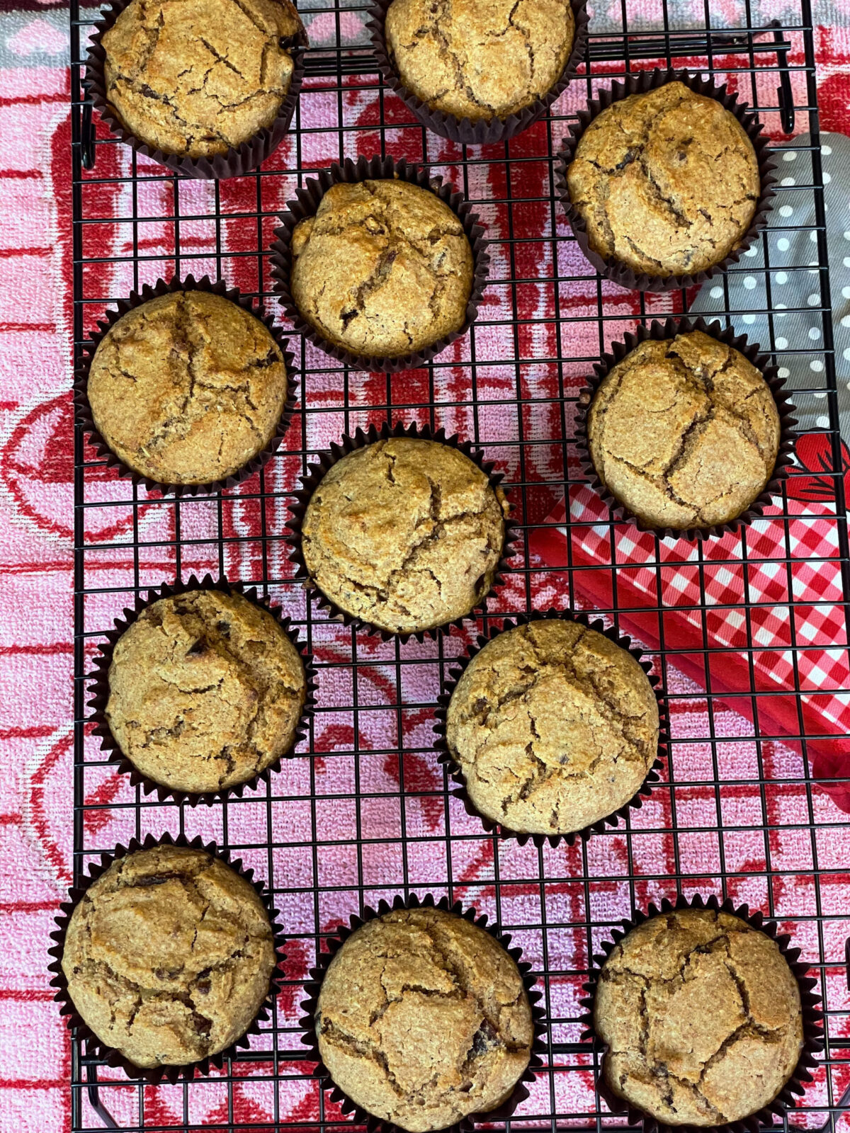 Bran muffins cooling on a cooling wire rack, with pink tea towel underneath and  red and white oven mitt.