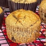 A close up of a bran muffin on a cooling rack with the paper case removed, more muffins with brown paper cases in background.