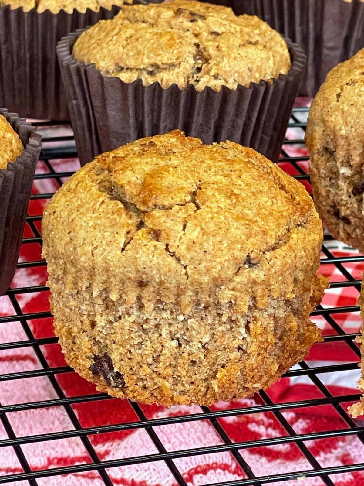 A close up of a bran muffin on a cooling rack with the paper case removed, more muffins with brown paper cases in background.