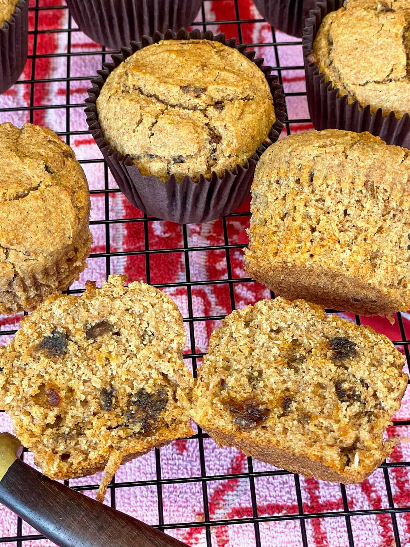 Bran muffins cooling on wire rack, with one halved down the middle and brown handled knife to side, pink tea towel underneath.