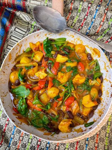 Vegan jalfrezi curry in Wok with silver vintage serving spoon to side, and colourful patterned table cloth background.