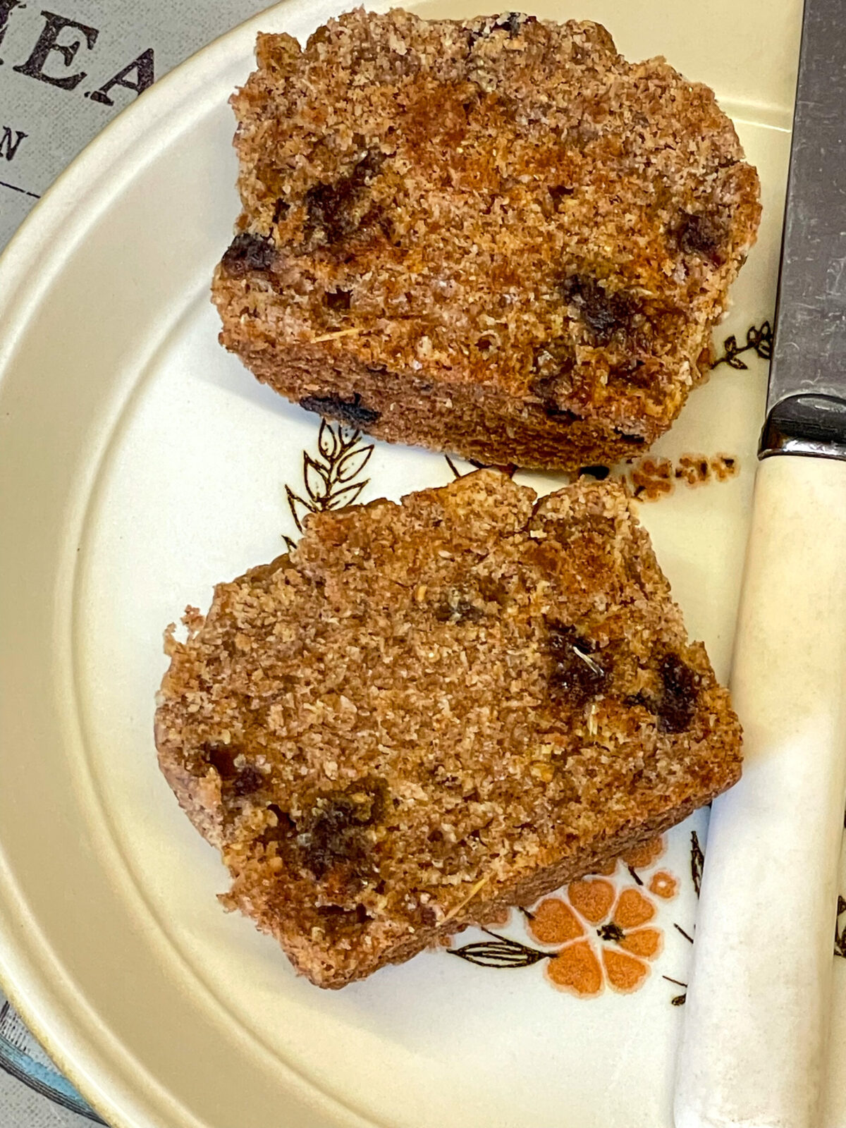 Bran muffin that has been sliced in half and toasted, on a small flower imaged plate with cream handled butter knife.