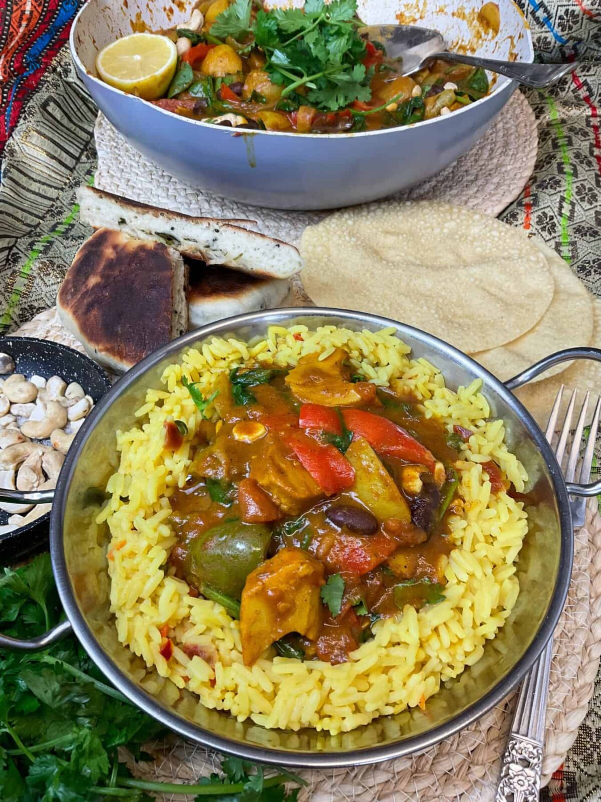 A single silver bowl of curry and rice, with Wok in background with curry and cilantro garnish, more cilantro to sides along with naan bread and poppadoms, and toasted cashews.