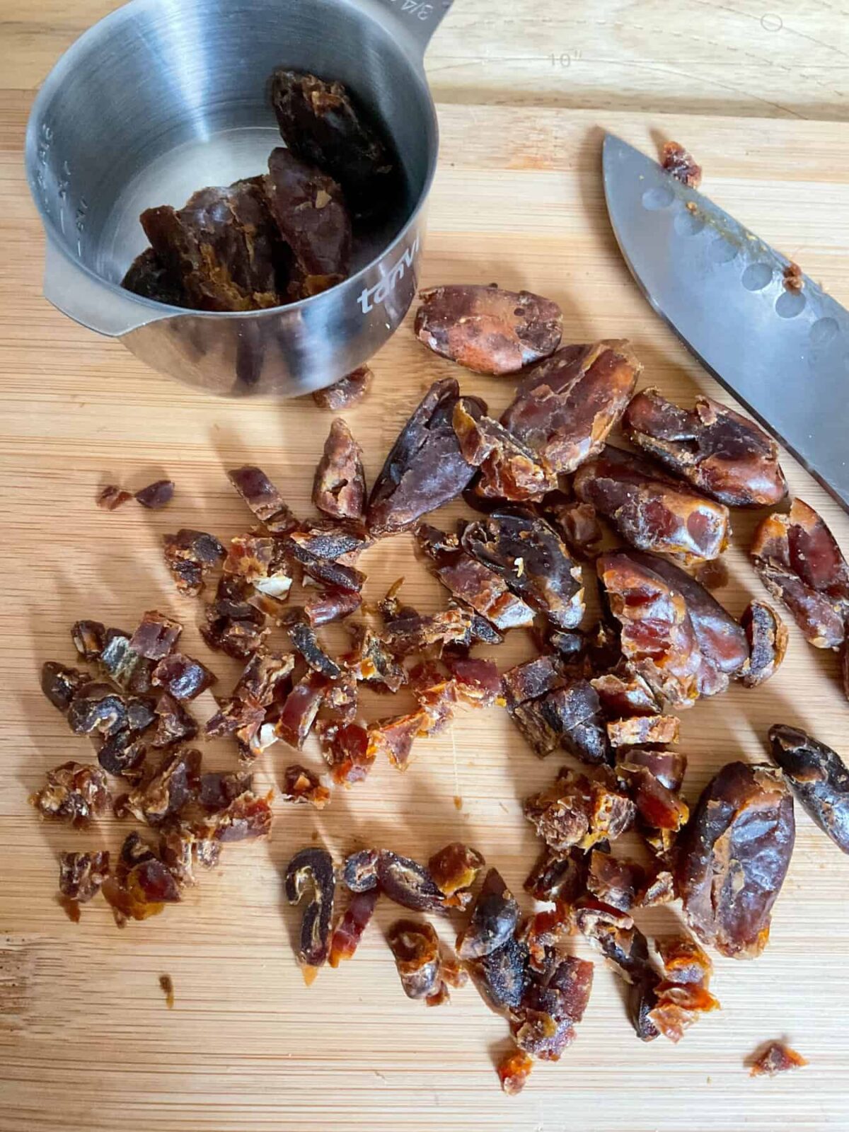 Dates chopped on a wooden chopping board, with measuring cup and silver knife to side.