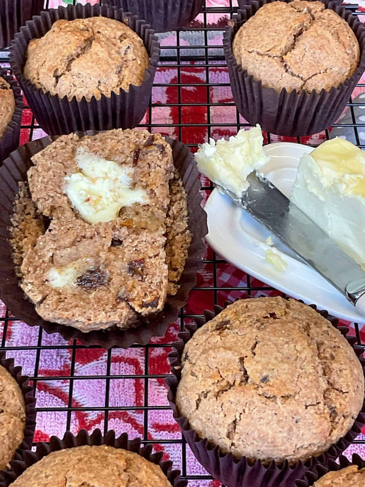 Bran muffins on wire rack with one split open and vegan butter spread over, small butter tub with butter knife to side.