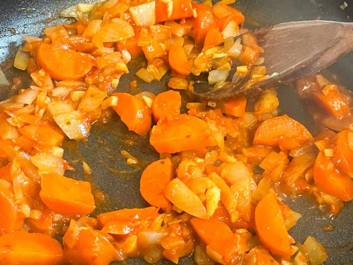 Vegetables sauteing with red Thai curry paste in skillet pan with wooden spoon.