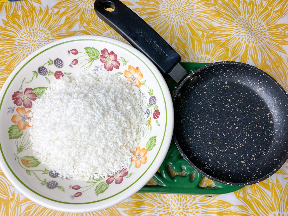 A bowl of desiccated coconut with a small fry pan sitting on a green trivet with a yellow flower table cloth.