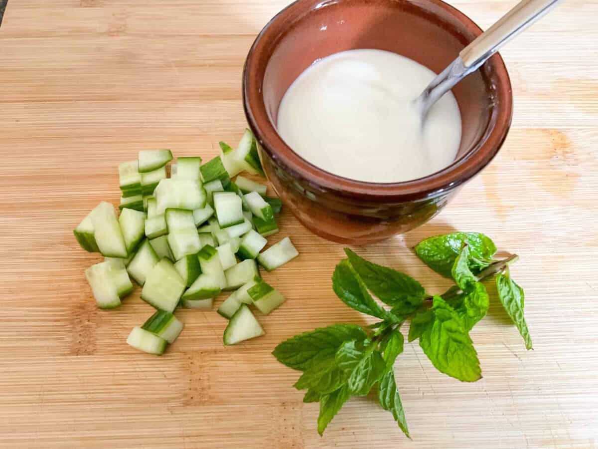 Wooden cutting board with small brown dish filled with yogurt, chopped cucumber and fresh mint leaves.