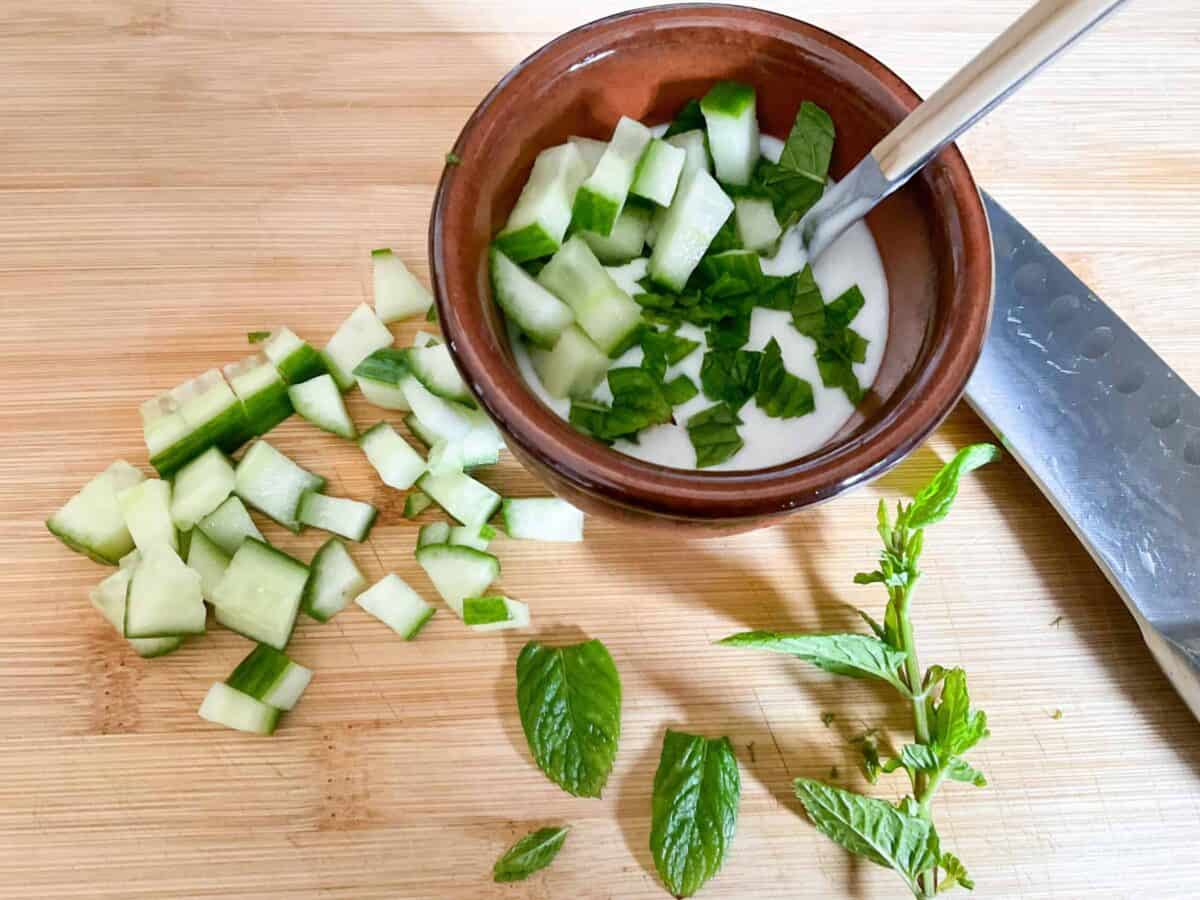 Diced cucumber and shredded mint leaves added to yogurt in brown pot.