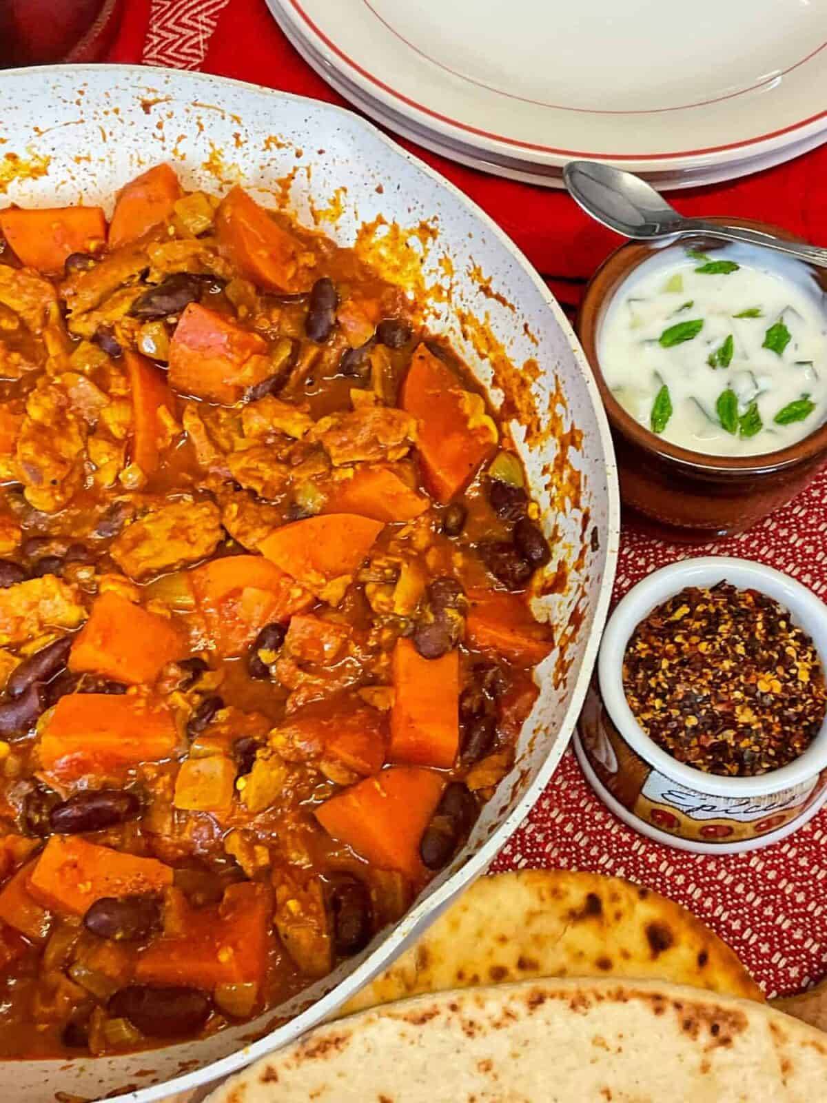 Side view of curry in cooking pan, with small dish of chilli flakes to side and small pot of home-made raita to side, naan breads and empty plates to side, red stripe table cloth background.