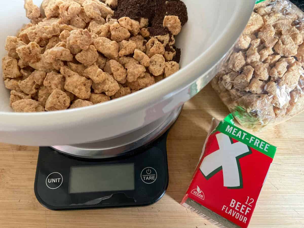 Soya chunks added to bowl on scale, soya chunk package to side, and tub of vegan stock cubes on cutting board.