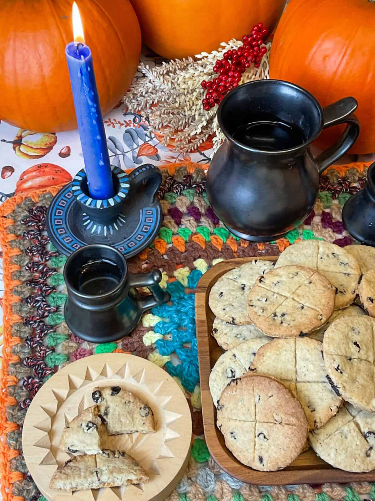 Blue candle lit beside pile of soul cakes and small wooden circle plate with a broken pieces of soul cake, three pumpkins in background with reeds and berry display.