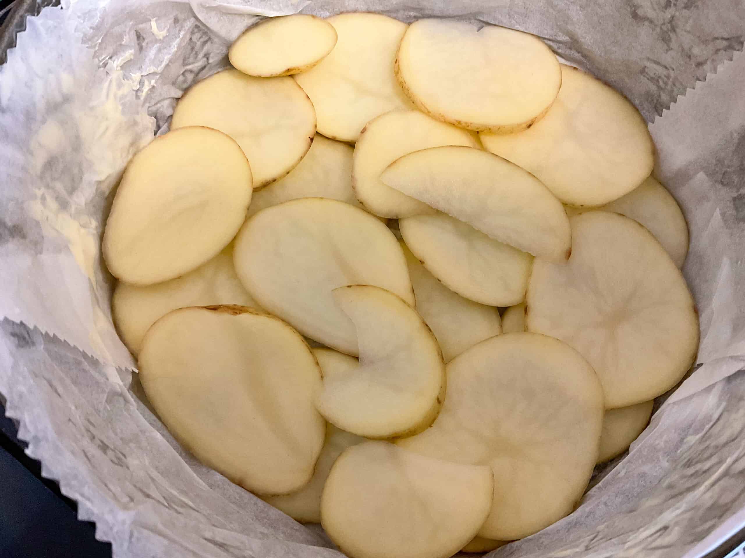 A layer of thin sliced potatoes at bottom of a parchment paper lined cake tin.