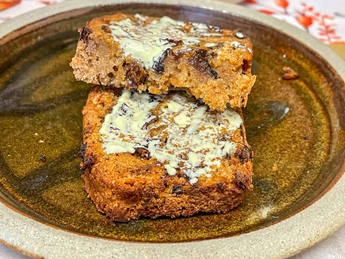 Slice of fruit loaf from the air fryer spread with butter and on a dark brown rustic plate.