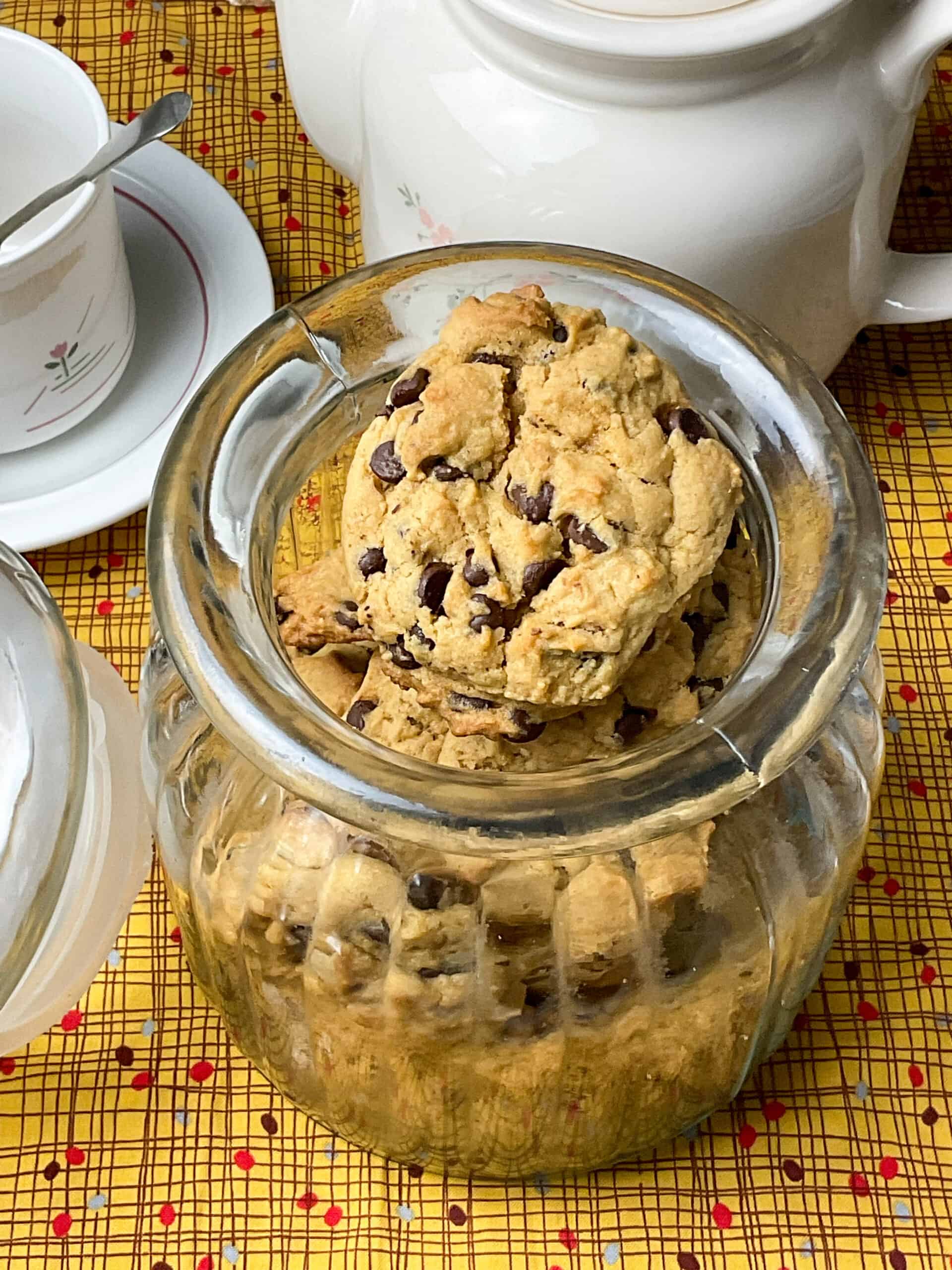 A glass cookie jar filled with chocolate chip cookies, tea pot and cup and saucer to side, and a yellow dotty patterned tablecloth.