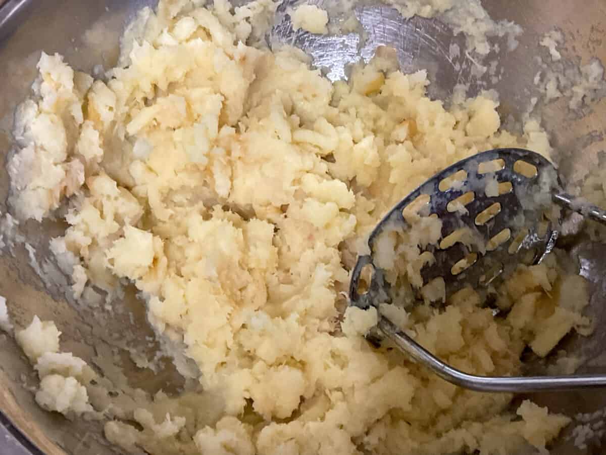 Parsnip chunks in a metal bowl being mashed with a potato masher.