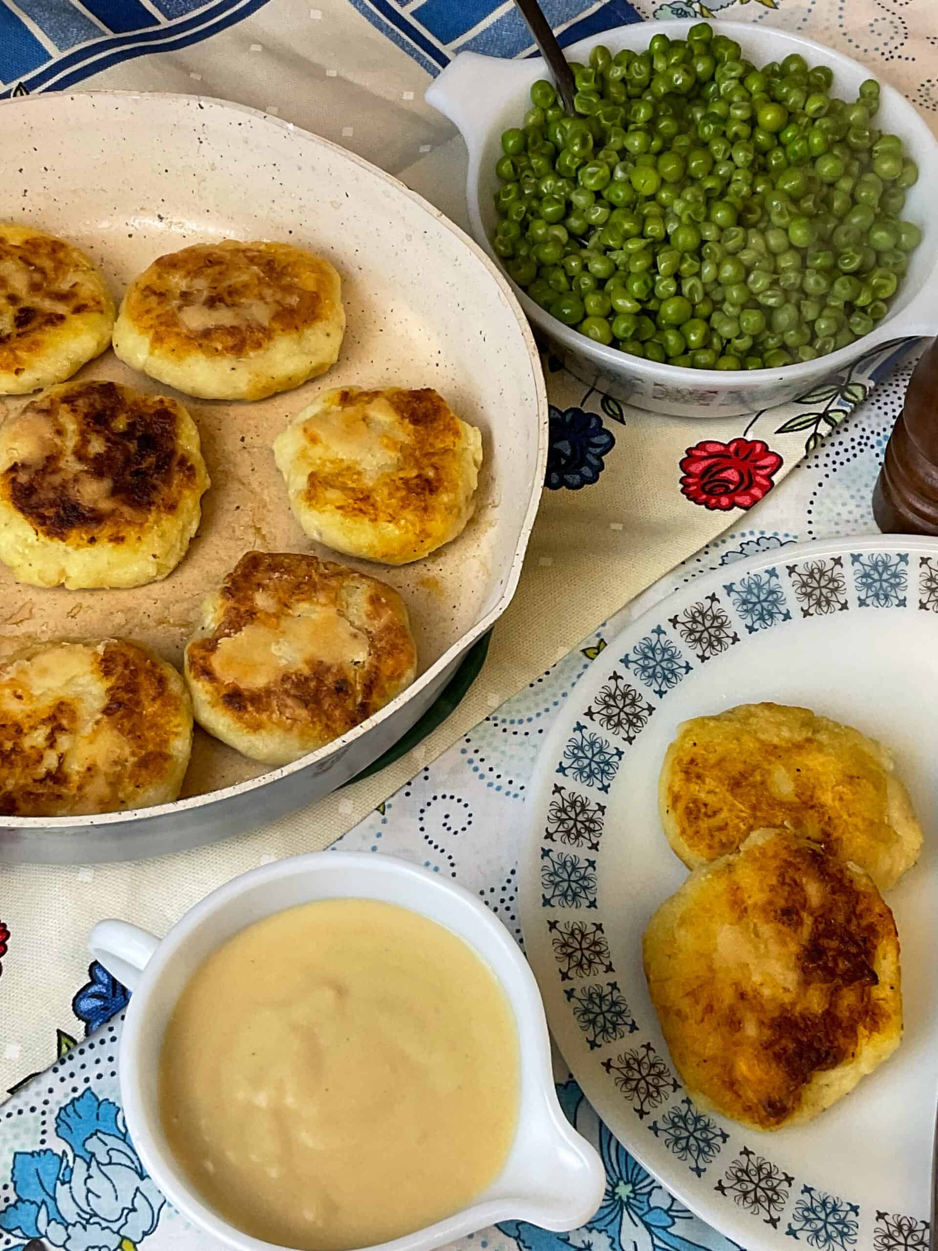 A fry pan with cooked parsnip patties and a small dish of peas to side, and small jug of cheese sauce and a plate with a few patties, and a blue and white flower patterned table cloth.